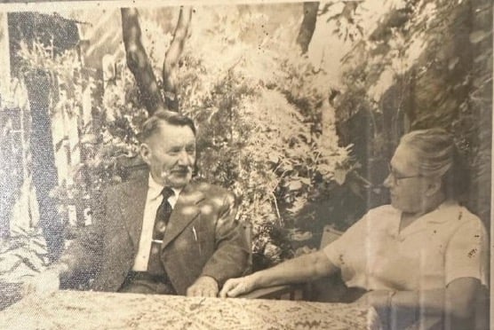 Foto antigua en sepia: un hombre de traje y una mujer conversan sentados a una mesa en un jardín.